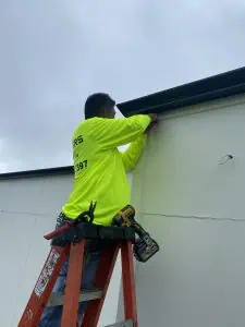 A person in a neon yellow shirt stands on an orange ladder, using a drill to attach something to the light-colored wall during a gutter installation. The overcast sky looms above, while another power tool rests beside them on the ladder.