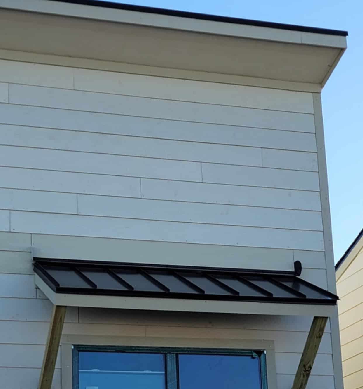 A modern building facade with light-colored wood siding showcases a sleek metal roof installation. A small overhang highlights the craftsmanship above a partially visible window, while the sky remains clear and blue.