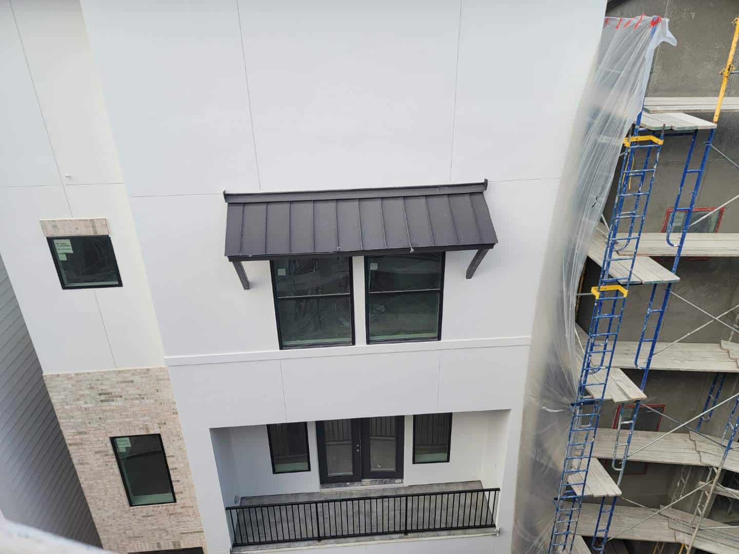 View of a multi-story building under construction. A black metal awning is above a pair of windows. A balcony with double doors is visible below. Scaffolding with blue and yellow beams, used for gutter installation, is to the right, covered by transparent sheeting.
