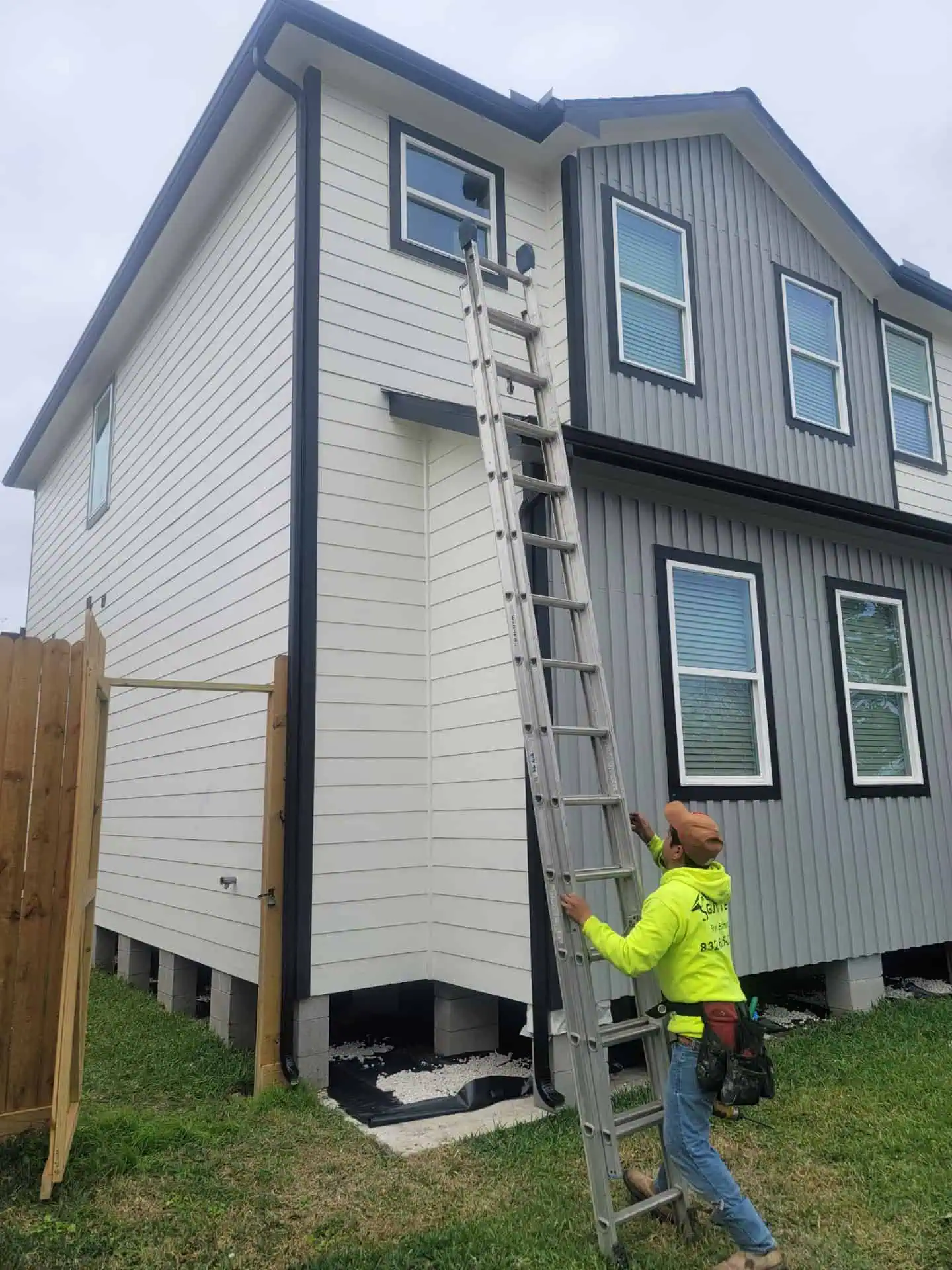 A worker in a neon shirt and hard hat stands at the base of an extended metal ladder, ready for gutter repair on a two-story house with gray and white siding. The home features multiple windows, while grass and a wooden fence complete the setting.