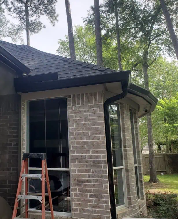 A brick house with a black gutter system in need of repair along the roof edge. A wooden ladder leans against the side, and tall trees are visible beyond the roofline. The windows reflect the greenery outside, subtly hinting at nature's influence on home design.
