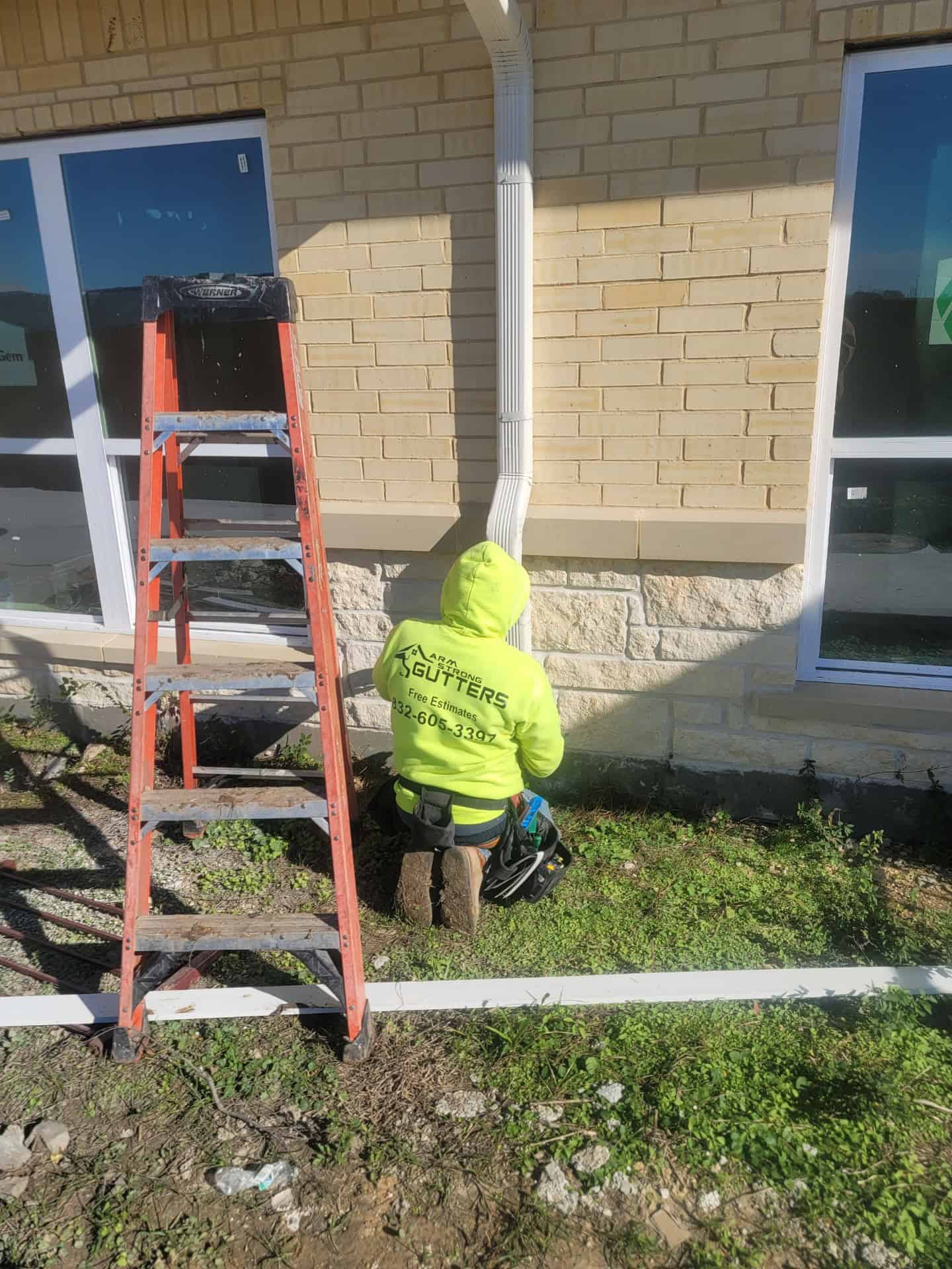 A worker in a bright yellow jacket kneels on the grass near a brick building, focused on gutter installation with care. A red ladder stands nearby as sunlight casts shadows on the wall and ground.