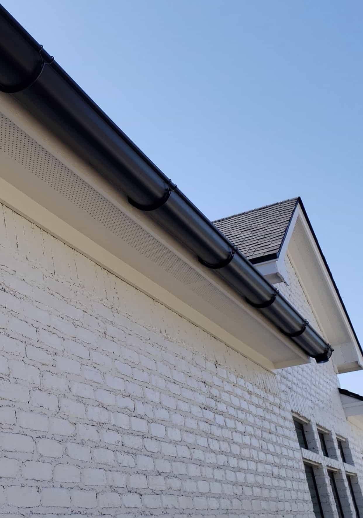 A section of a house's exterior features a white brick wall with pristine black gutters, showcasing expert gutter installation along the roofline. The dark shingled roof complements the scene, with a clear blue sky serving as the perfect backdrop.