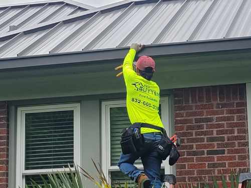 A person in a neon yellow shirt and red cap focuses on gutter repair atop the metal roof. As they lean against the brick building, their shirt displays a logo and contact number. Equipped with a tool belt and jeans, they work diligently above two visible windows.