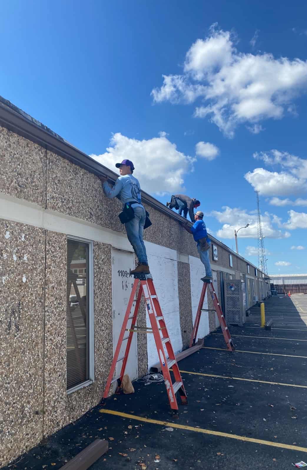 Three workers in blue shirts stand on ladders inspecting or repairing the roof of a single-story building on a sunny day. The team appears focused on a gutter installation task. The building has a textured façade, and a few fluffy clouds are visible in the blue sky.