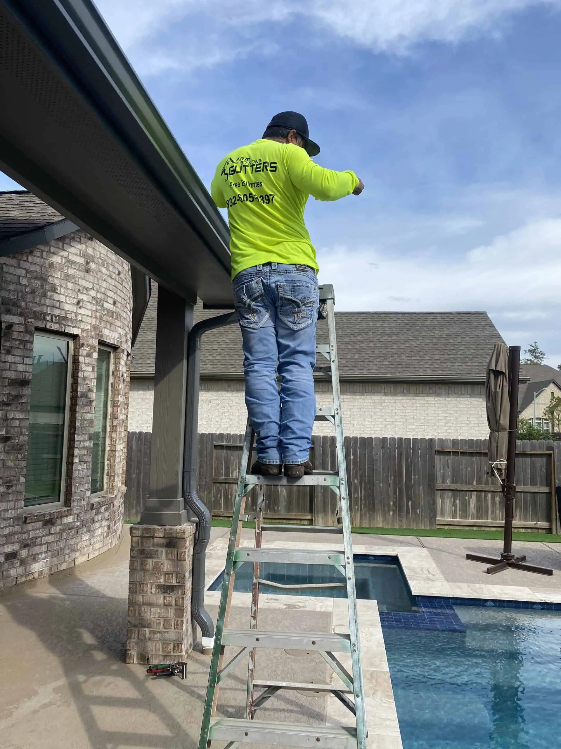 A person in a neon green shirt and jeans stands on a ladder, focusing on gutter installation next to a pool. The house is made of brick, with a wooden fence in the background under a partly cloudy sky.