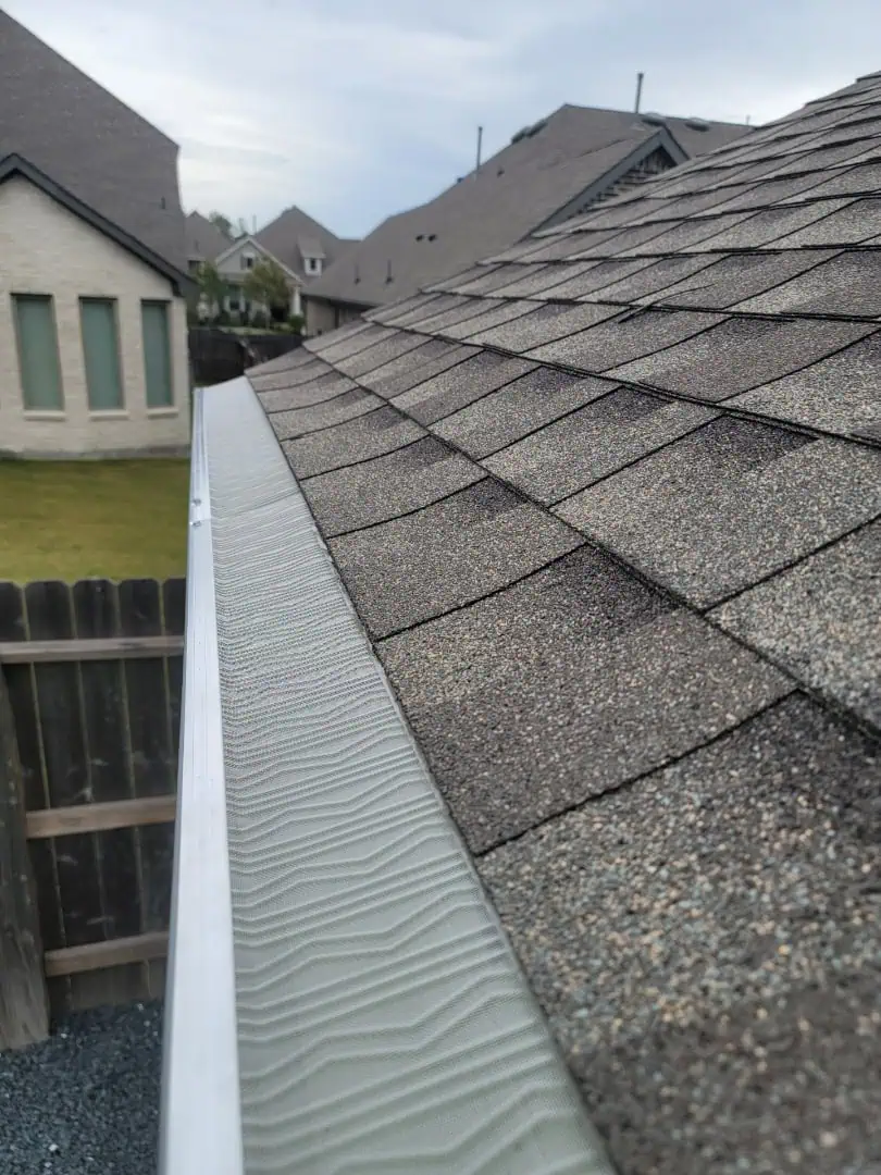 A close-up view of a house roof showcases textured asphalt shingles and a newly installed metal gutter guard, the result of expert gutter installation. The gutter runs along the roof's edge. In the background, similar houses with matching roofs stand under a cloudy sky.