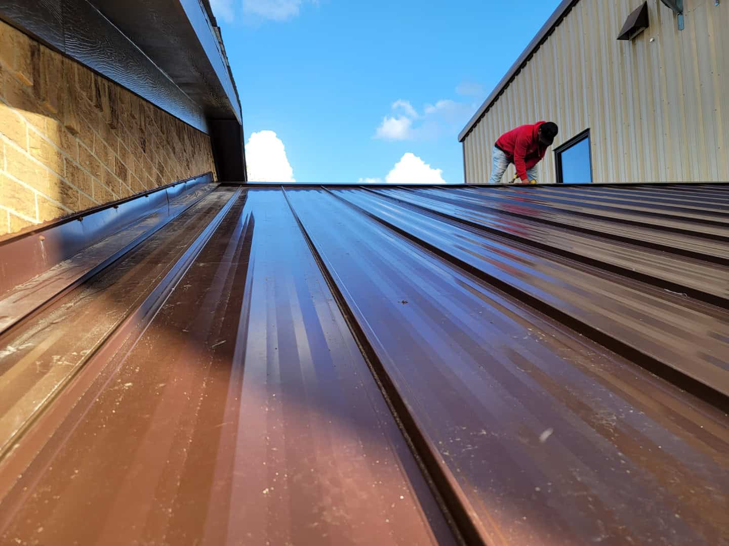 A person in a red shirt expertly handles metal roof installation on a sloped brown surface. The backdrop reveals a beige building under a blue sky with clouds, captured from the base of the roof, looking upward.