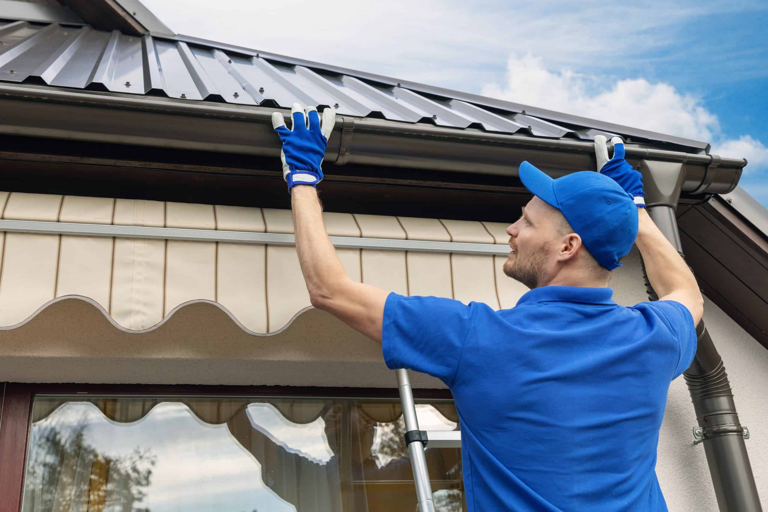 A man in a blue uniform and cap stands on a ladder, focused on gutter repair, as he inspects a metal roof gutter. Wearing blue gloves, he works diligently against the backdrop of a house with a large window and striped awning.
