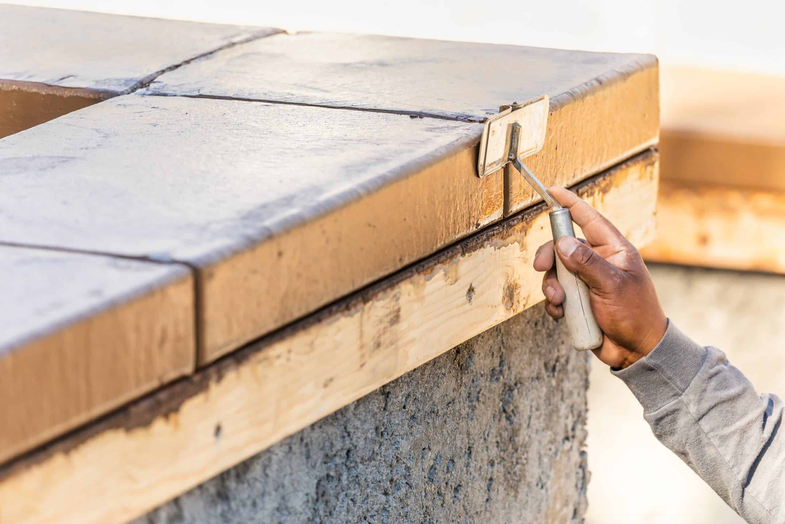A person skillfully smooths the edge of freshly poured concrete with a hand tool, next to a wooden form. The outdoor scene, under the bright sun, might be part of a larger project like gutter installation or similar tasks involving precision and craftsmanship.