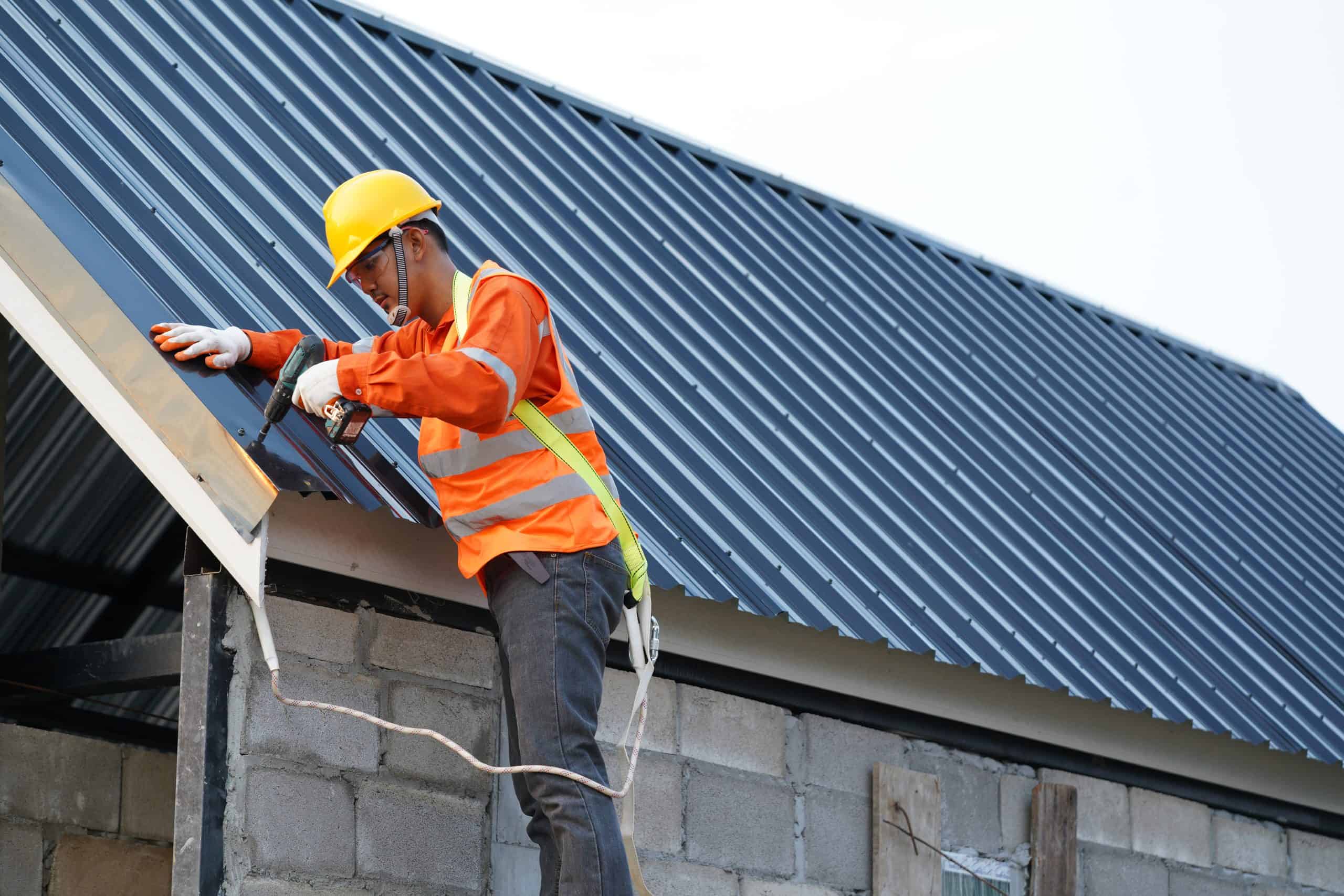 A construction worker wearing an orange safety vest and yellow hard hat is skillfully maneuvering a power tool on the edge of a metal roof, diligently focusing on coping cap installation while standing on a ladder attached to a brick wall.