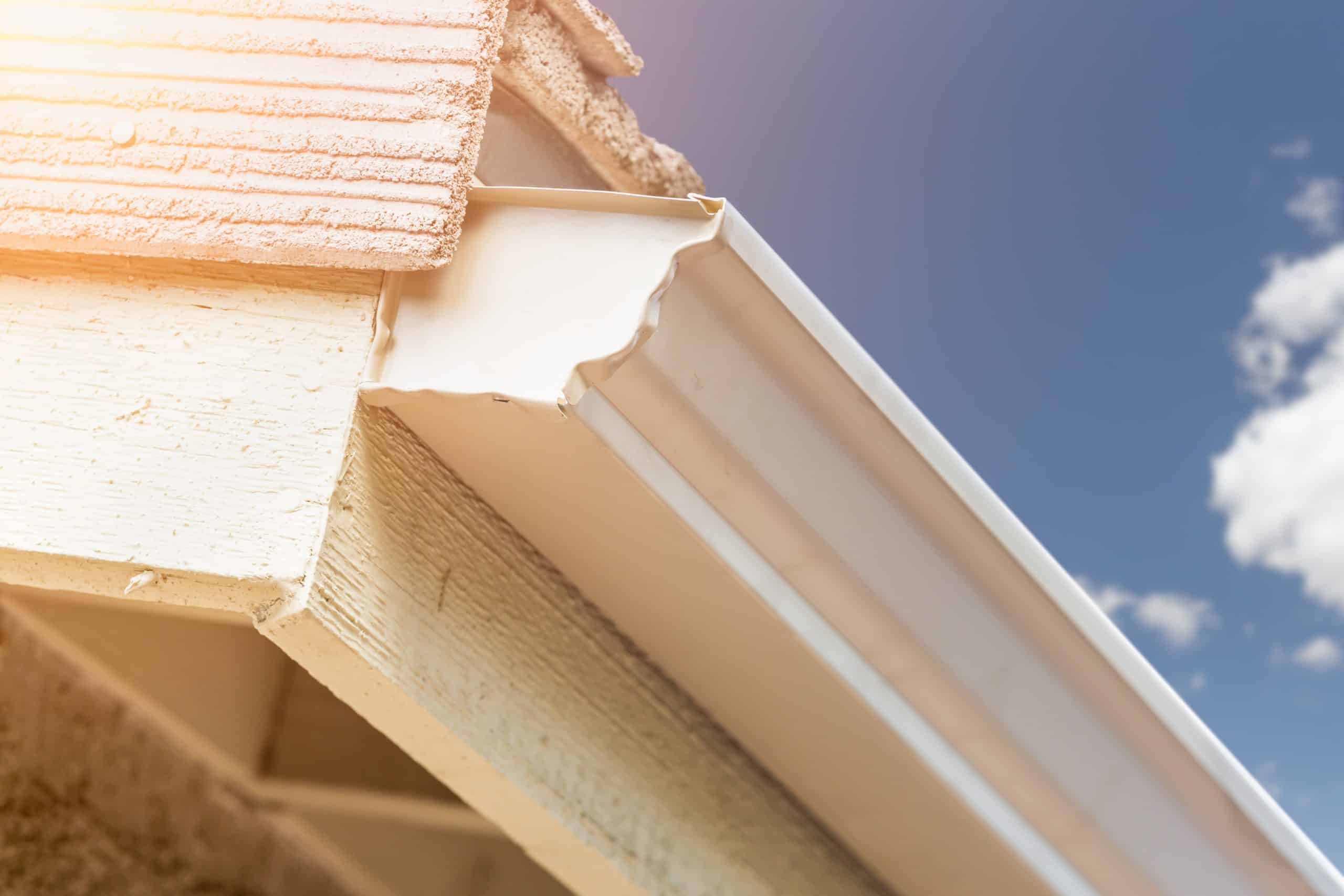 Close-up of a house roof corner featuring a beige gutter system under a textured, light brown roof. The sky is clear and blue with a few white clouds. Sunlight casts a warm glow on the structure, highlighting the recent gutter installation.