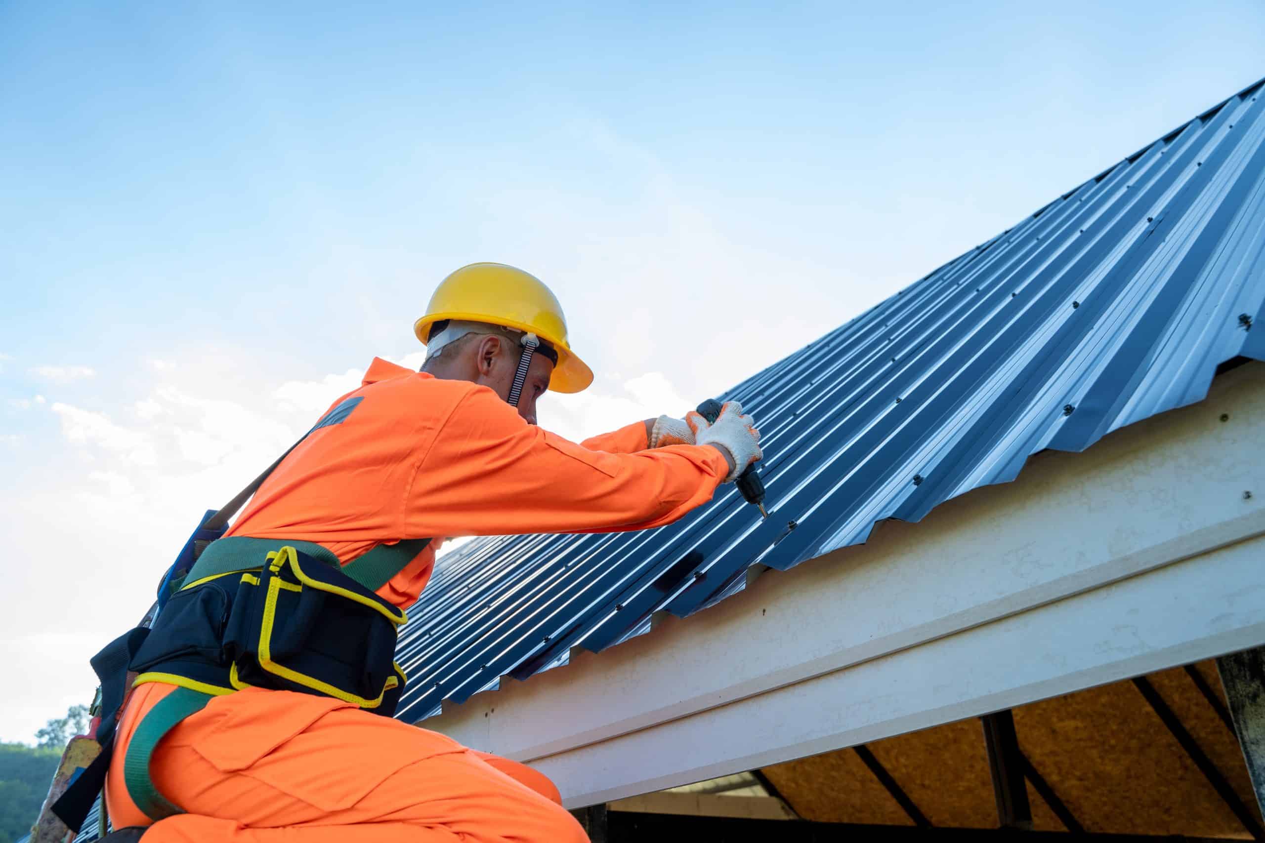 A construction worker in an orange safety suit and yellow helmet is securing a metal roof with a drill, expertly managing the metal roof installation. The worker is on a white structure against a clear, blue sky.
