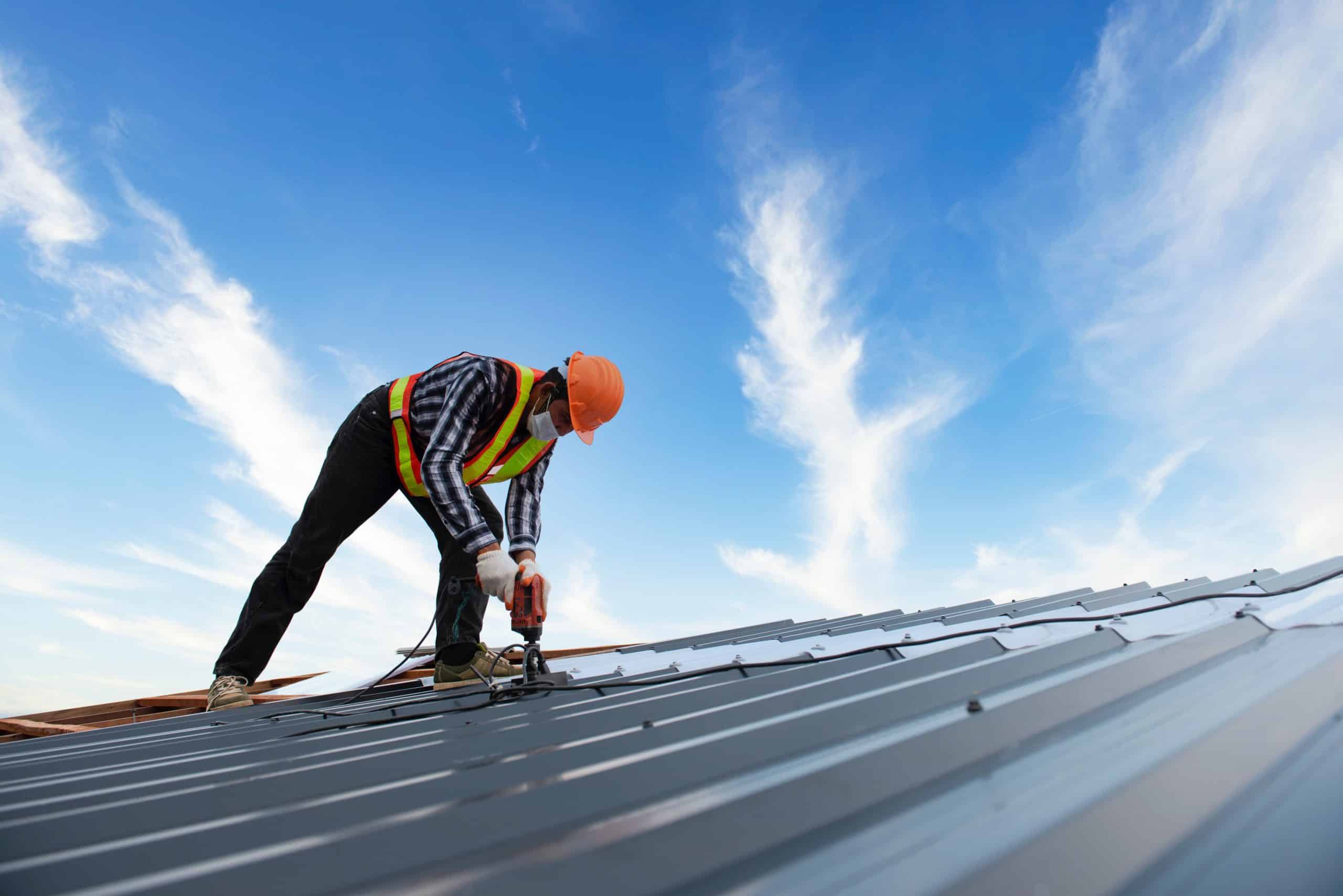 A construction worker in a safety vest and helmet uses a power drill for metal roof installation, securing sheets on a rooftop. The sky is clear with wispy clouds, emphasizing the bright daylight.