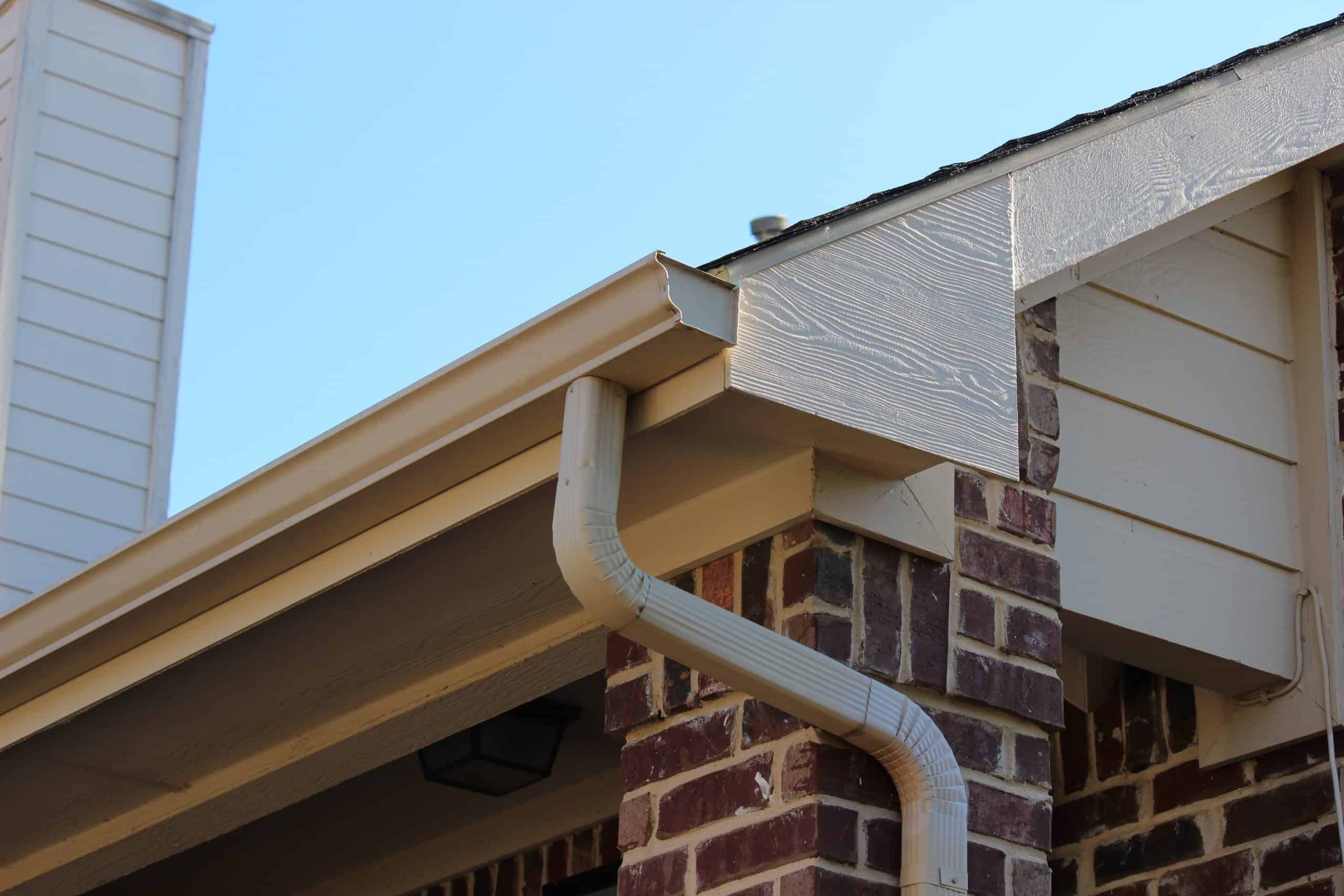 Close-up of a house corner with beige gutters and downspout attached to the roof, expertly showcasing recent gutter installation. The house features brick walls and white siding under a clear, blue sky.