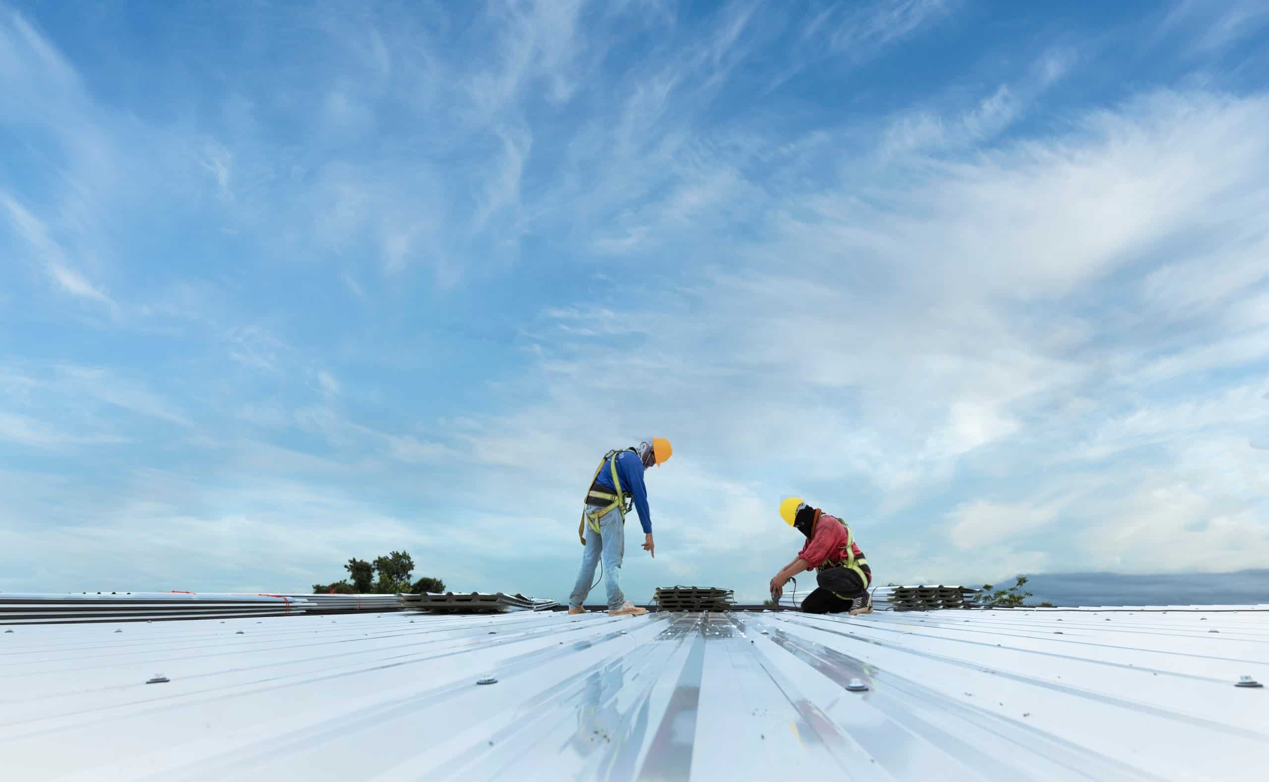 Two construction workers in safety gear are busy with metal roof installation under a blue sky with scattered clouds. One stands pointing at the roof while the other kneels, expertly handling tools and materials to ensure everything is securely in place.