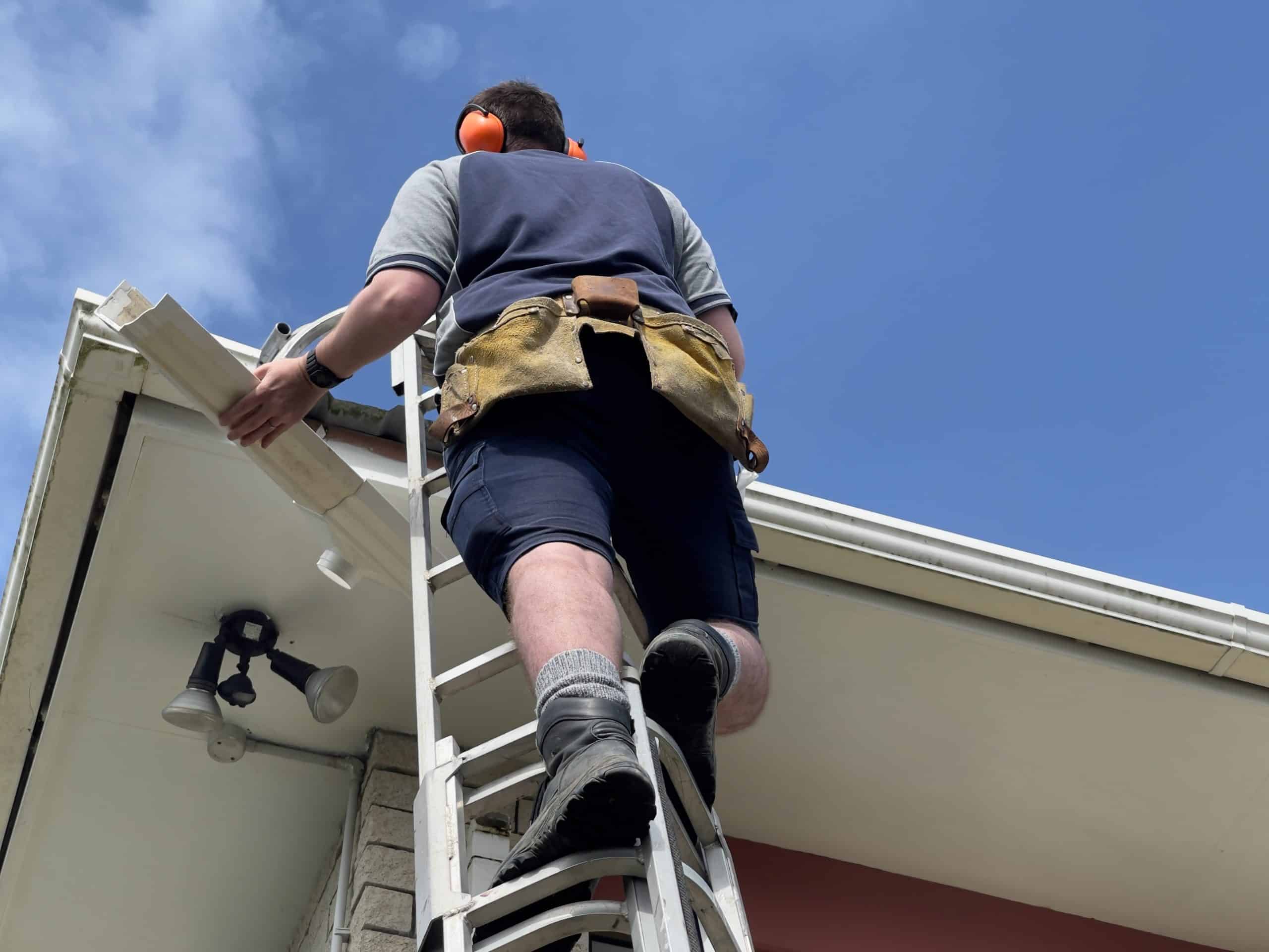 A person wearing a tool belt and ear protection is on a ladder, fixing a security camera under the eaves of a house, right next to where they recently completed gutter repair. The sky is clear and blue.