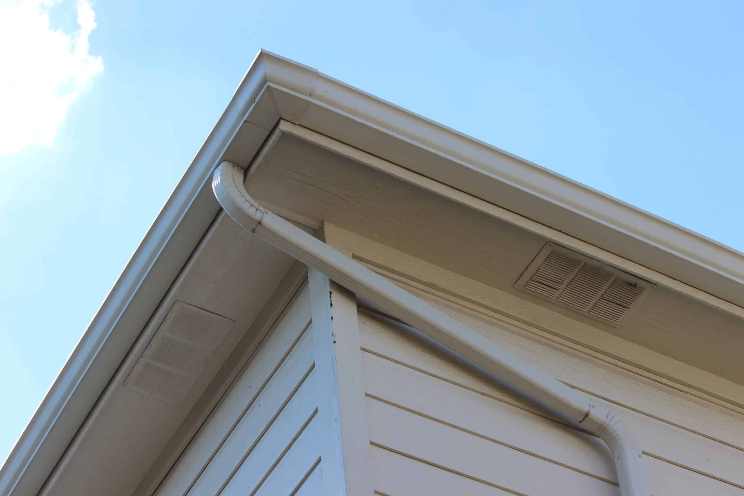 View from below of a house roof corner showing a white downspout extending from the gutter down the side. The sky is clear and blue, highlighting the modern and neat design of the building's exterior, with recent gutter repair enhancing its sleek appearance.