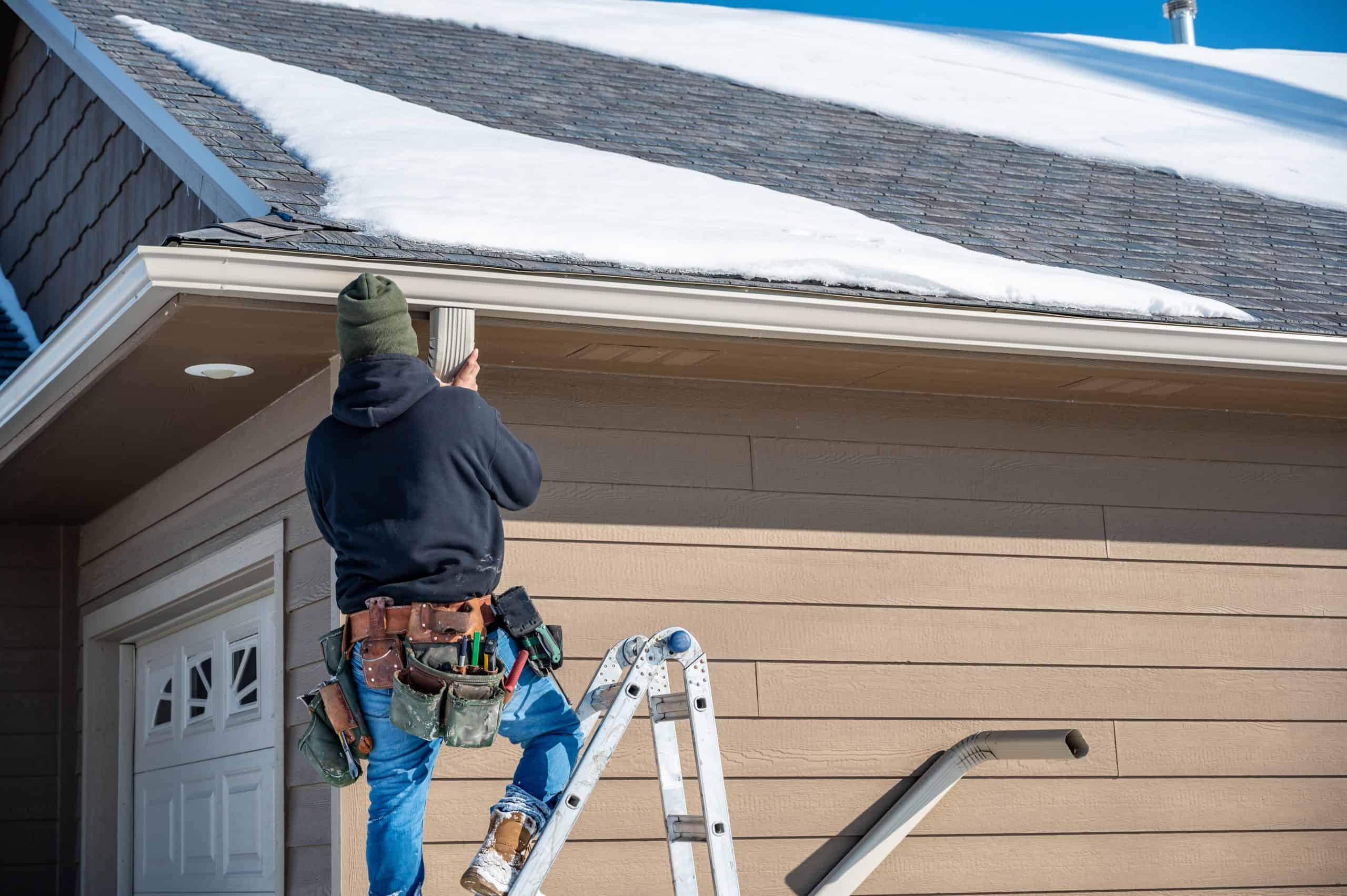 A person in a black hoodie and jeans stands on a ladder, focused on gutter repair on the snowy house roof. Equipped with a tool belt, they ensure everything is secure under the clear blue sky.