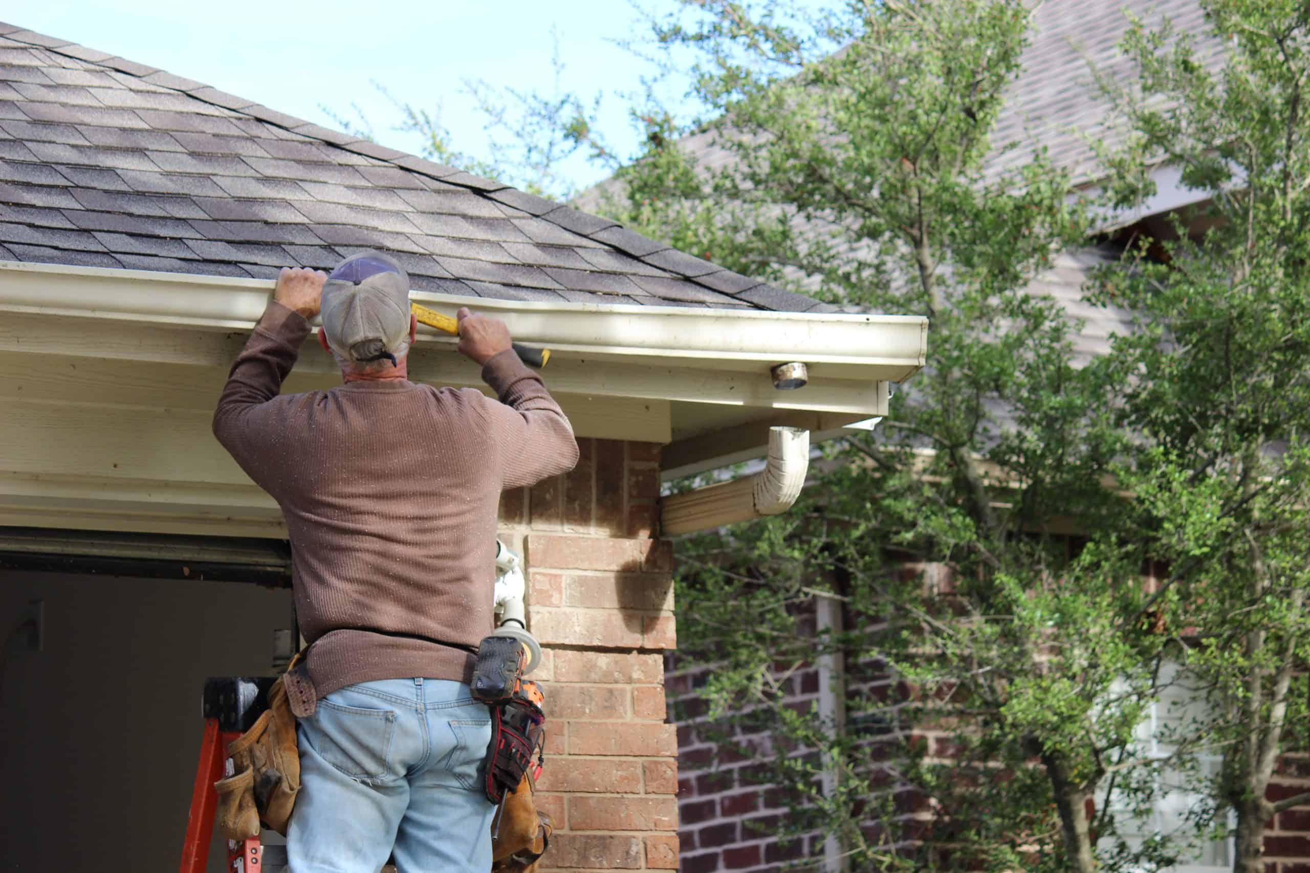 A person wearing a cap and tool belt stands on a ladder, using a yellow level to check the alignment of a gutter during its installation on a brick house. Greenery is visible in the background.
