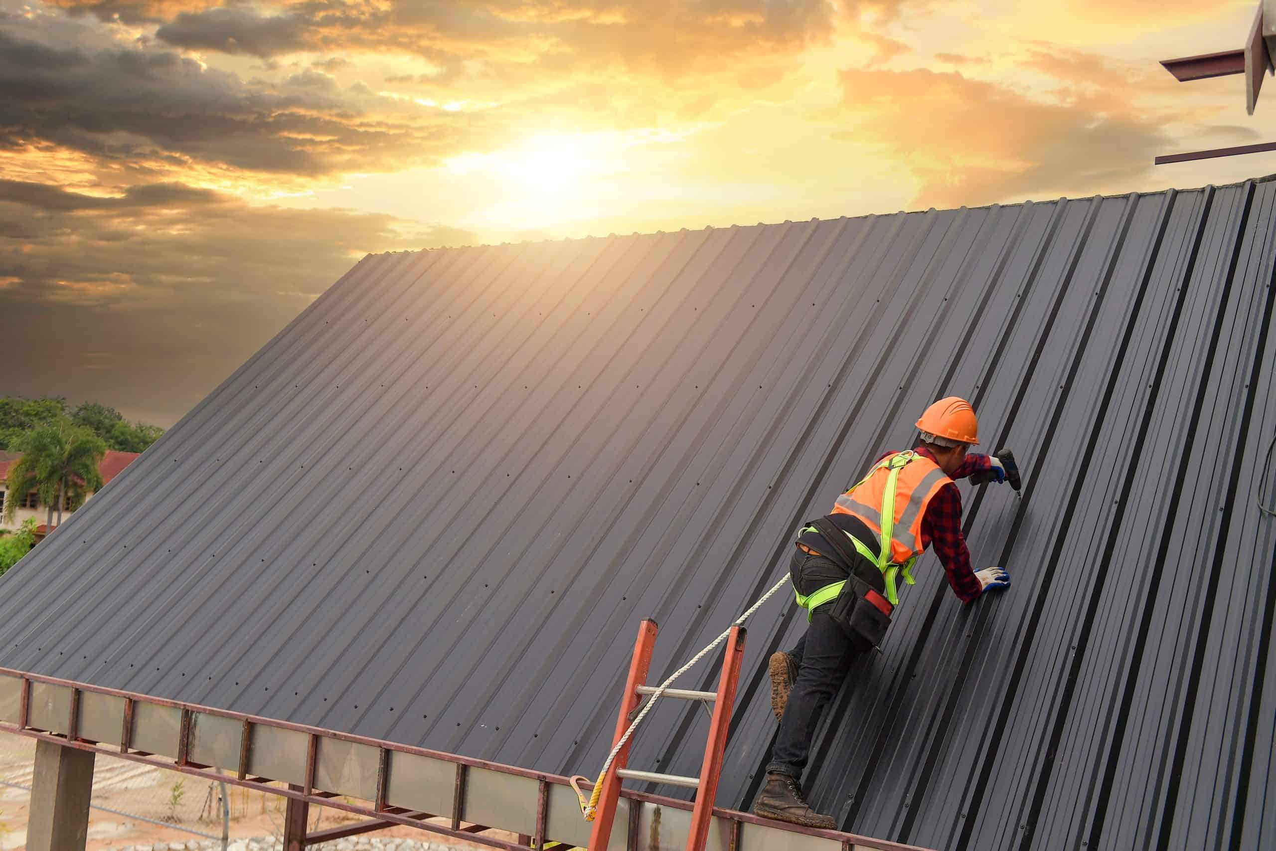 A construction worker wearing a safety vest and helmet is climbing up a ladder for metal roof installation. The sun sets in the background, casting a vibrant glow over the dark gray surface and clouds.