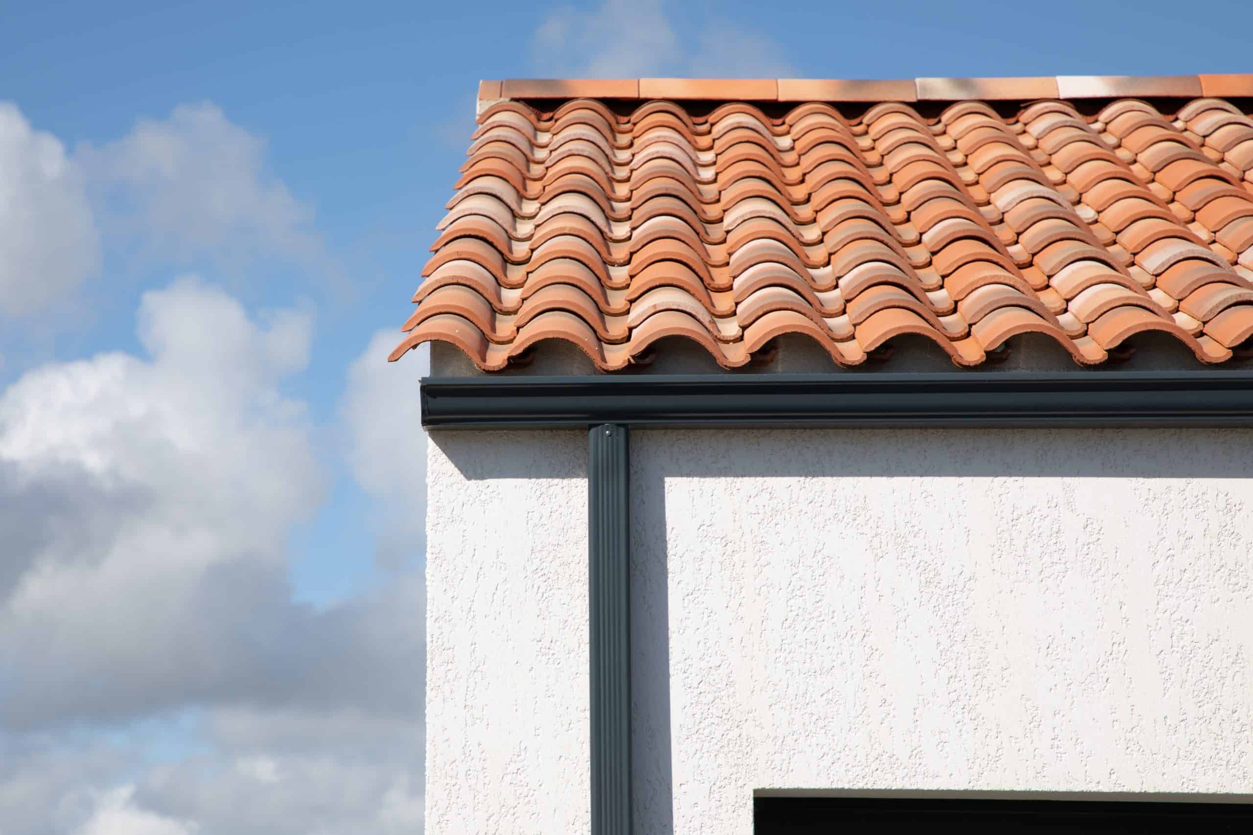 A house with white stucco walls and a red clay tile roof featuring a classic wavy pattern stands against a blue sky with scattered clouds. The recent gutter repair enhances its charm, ensuring rainwater flows smoothly around the picturesque structure.