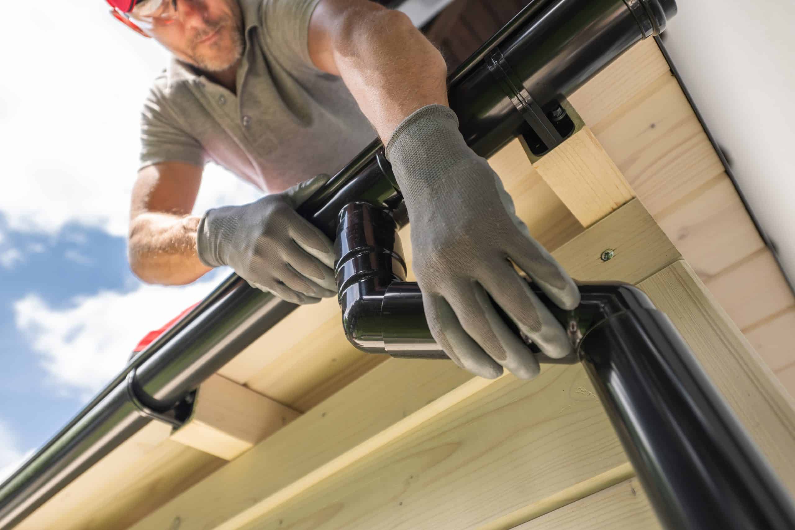 A person wearing gloves and protective eyewear is focused on gutter installation, fitting black plastic gutters securely onto a wooden roof structure under a clear sky.