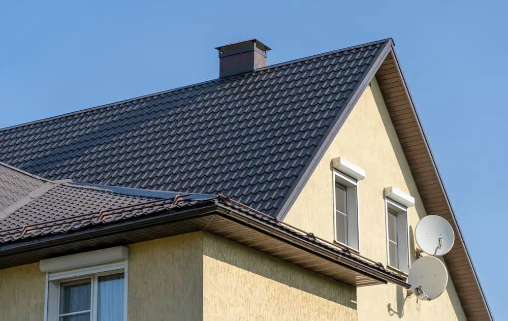 A yellow house with a steep, dark gray metal roof and a chimney boasts newly installed gutters. Two satellite dishes are attached to the side, while several windows reflect the clear blue sky.