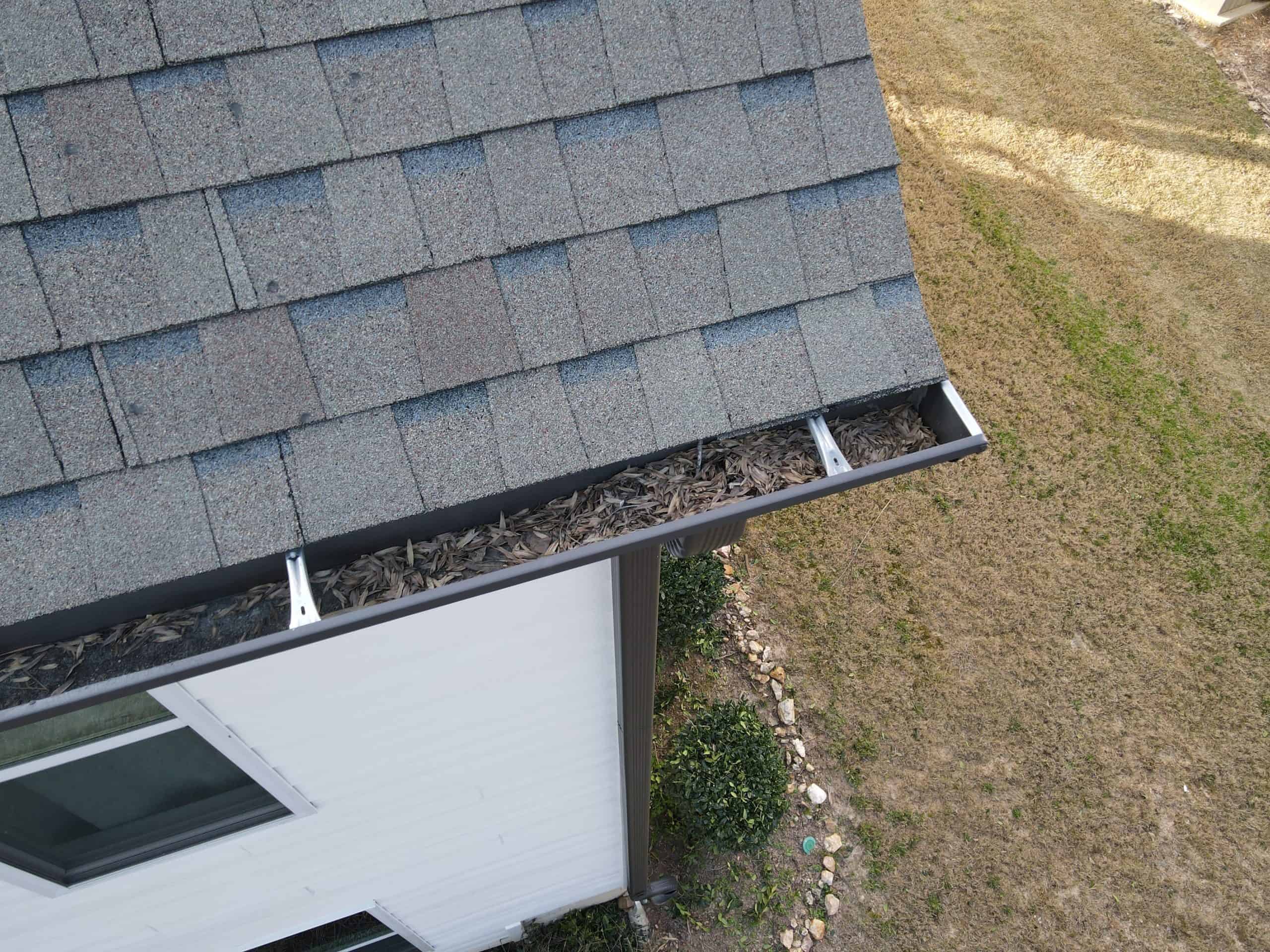 Aerial view of a house gutter filled with dry leaves and debris, in need of gutter installation. The roof is covered with gray shingles, the house has white siding, and surrounding it is a grass lawn with scattered stones and shrubs.