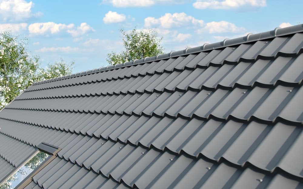 Gray metal roof with tile pattern under a blue sky with scattered clouds, showcasing expert metal roof installation. Two trees are partially visible in the background.