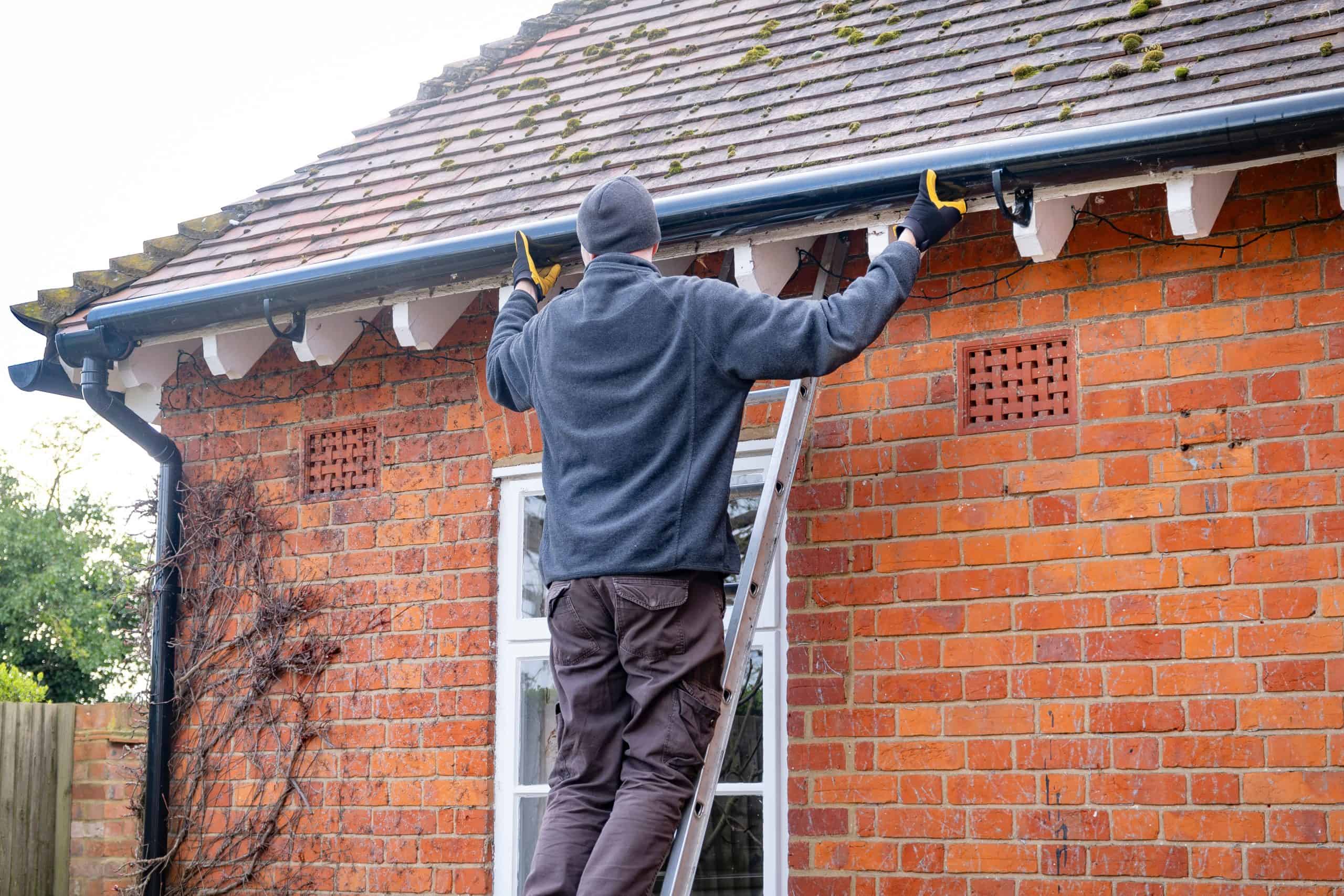 A person wearing a dark beanie and sweater stands on a ladder, engaged in gutter repair on a brick house. The roof, touched by moss, gleams under the clear day's light.