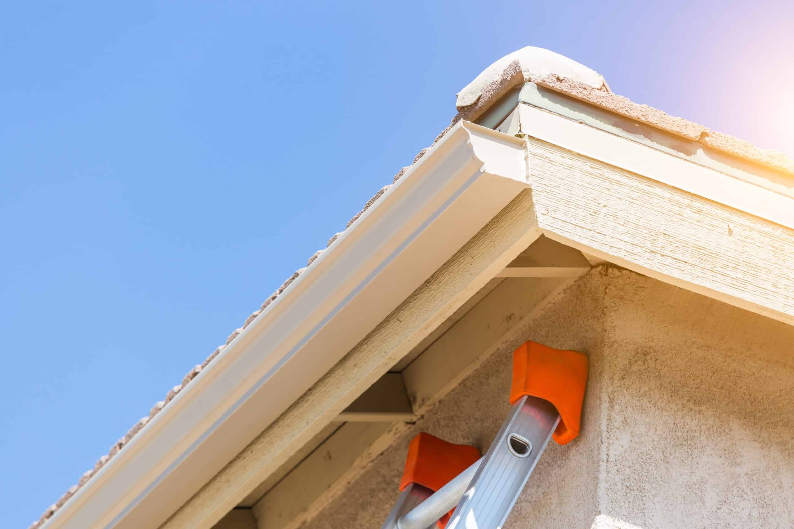 A close-up view of a building's corner roof edge with rain gutters under a clear blue sky hints at recent gutter installation. A ladder with orange padding leans against the house, suggesting maintenance or cleaning work. Sunlight gently illuminates the scene.