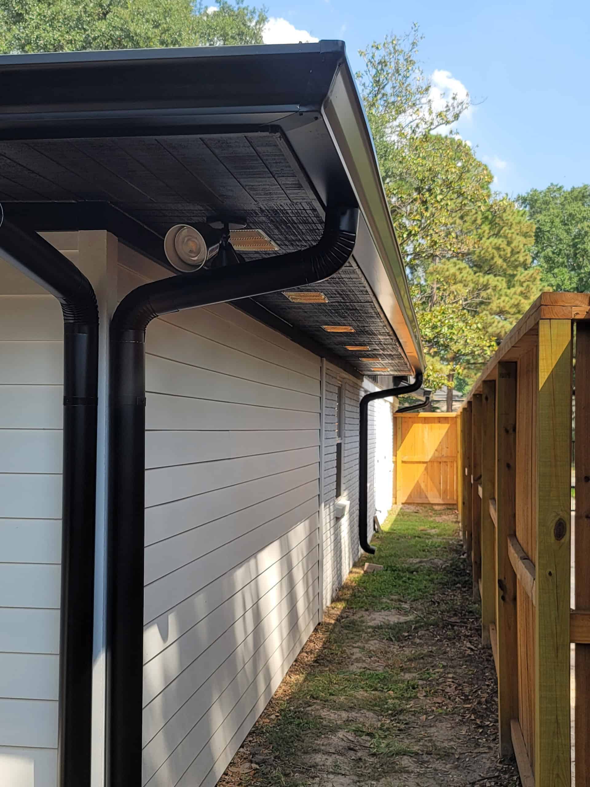 Side view of a house with white siding, a black gutter system, and a wooden fence on the right. The gutter extends along the roofline with downspouts attached. Trees and a clear sky can be seen in the background, showcasing expert metal roofing in Harris County, TX.