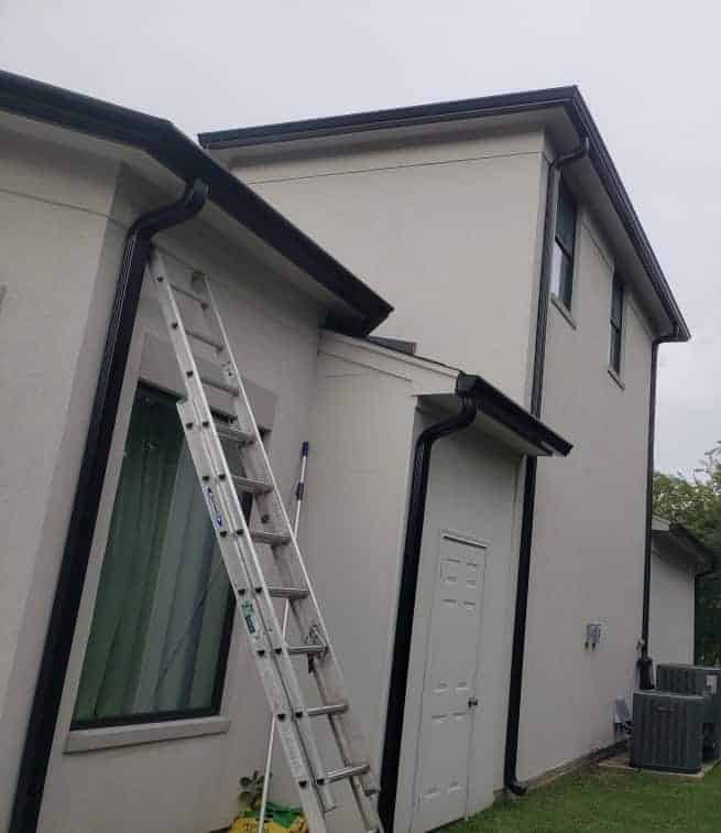 A tall ladder leans against the side of a two-story beige house with black trim. The house, possibly serviced by Gutter Services Harris County TX, features multiple windows and a white door, surrounded by grassy lawn and trees. An air conditioning unit is visible at the back.