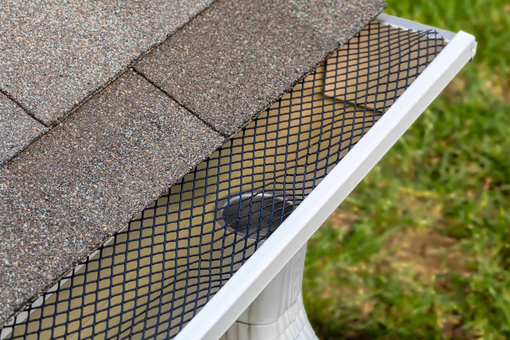Close-up of a house roof with asphalt shingles, a white rain gutter, and a black mesh gutter guard covering the gutter to prevent debris from entering. Green grass is visible in the background.