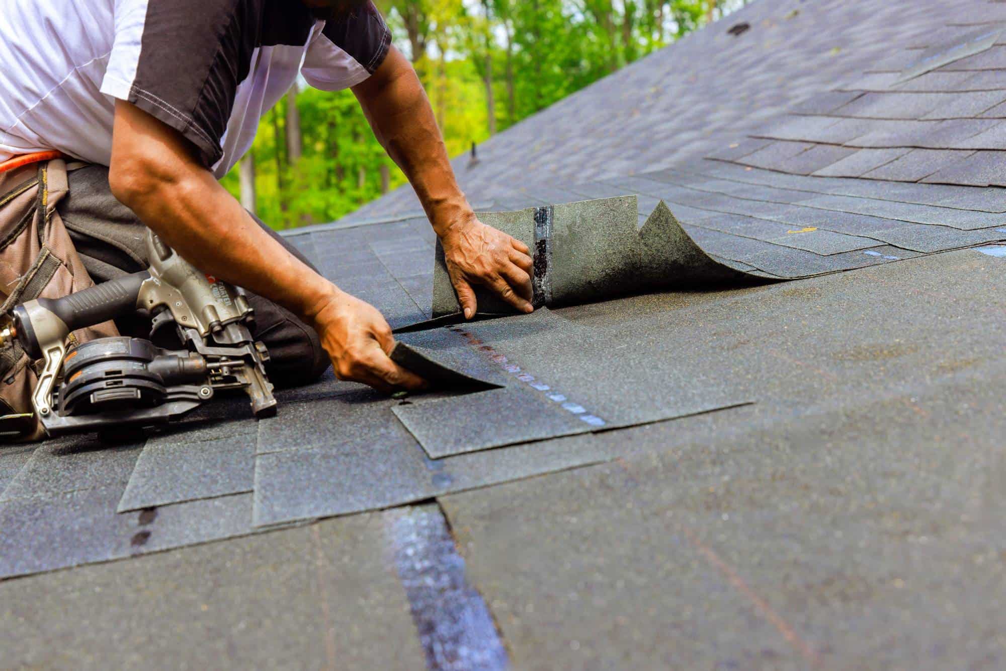 A person installs asphalt shingles on a sloped roof, using tools and aligning the shingles carefully. Trees and greenery are visible in the blurred background.
