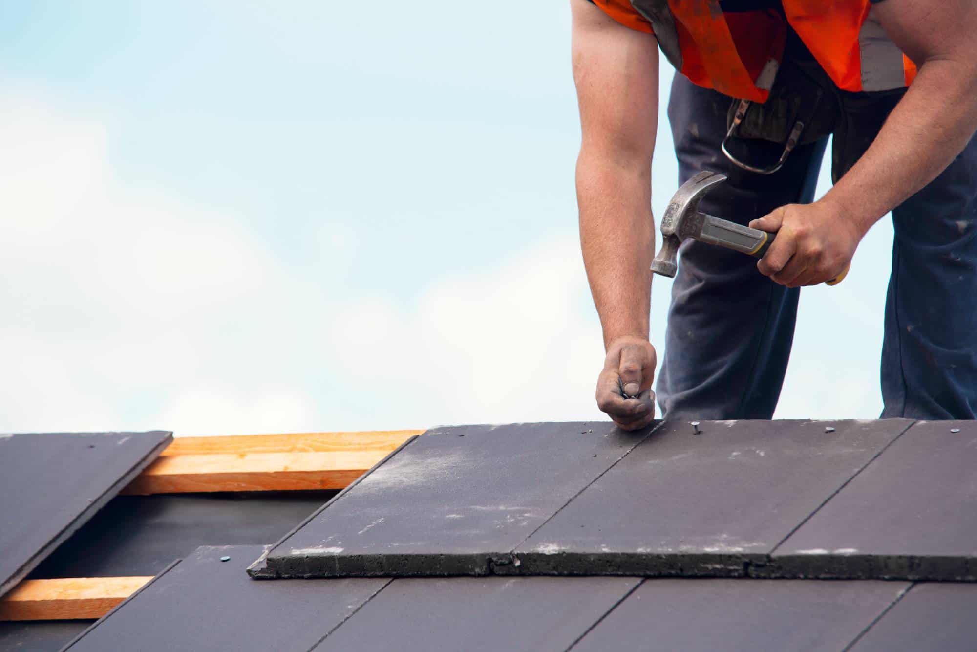 A roofer wearing an orange safety vest hammers a nail into dark grey roof tiles against a cloudy sky.