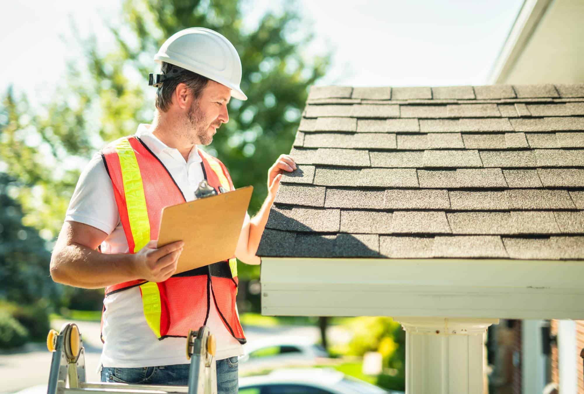 A man wearing a white hard hat and orange safety vest stands on a ladder, holding a clipboard and inspecting the shingles on a house roof in bright daylight.