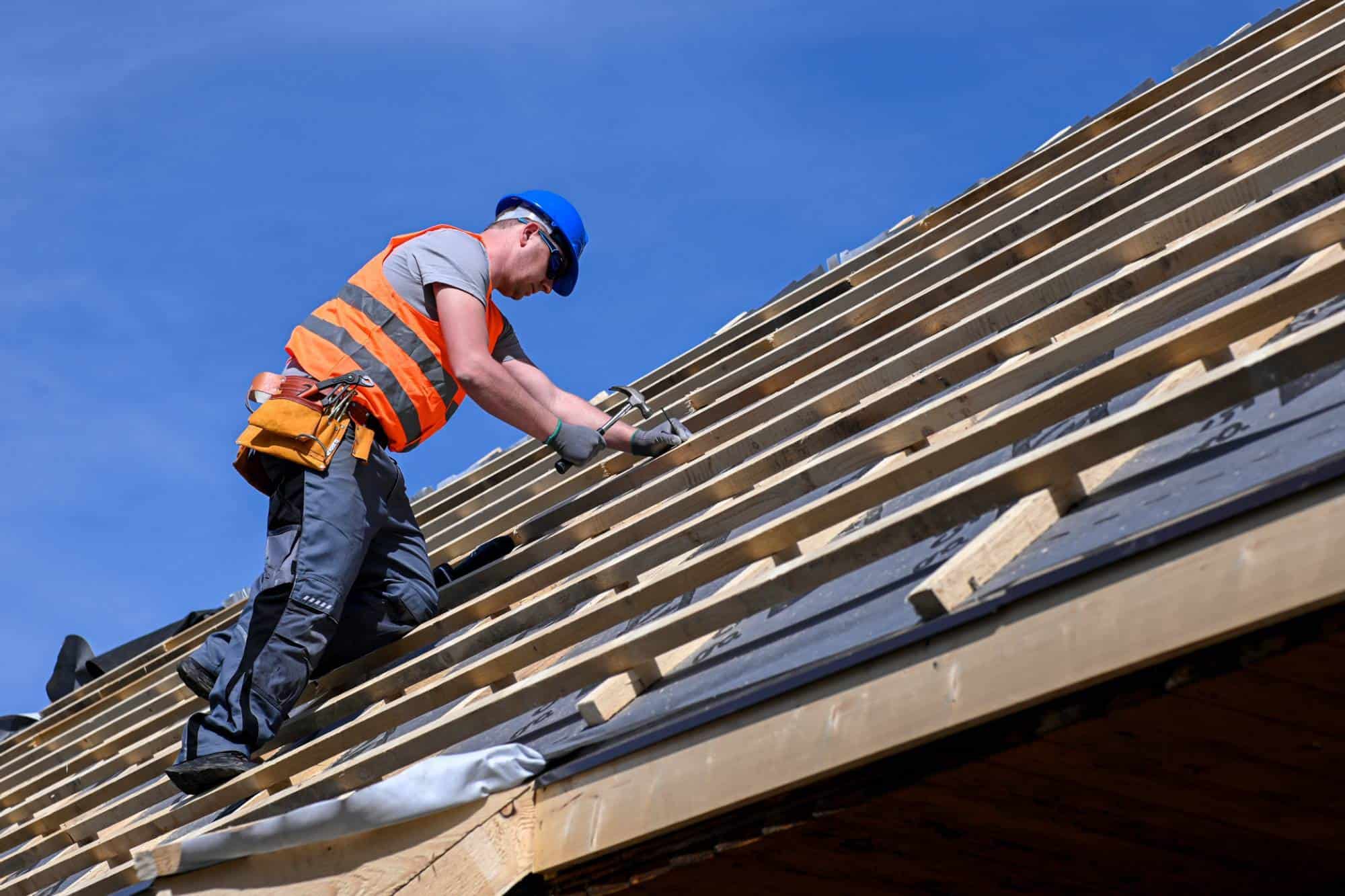 A roofer wearing a blue hard hat and orange safety vest kneels on a roof under construction, using a hammer to nail down materials onto the wooden battens under a clear blue sky.