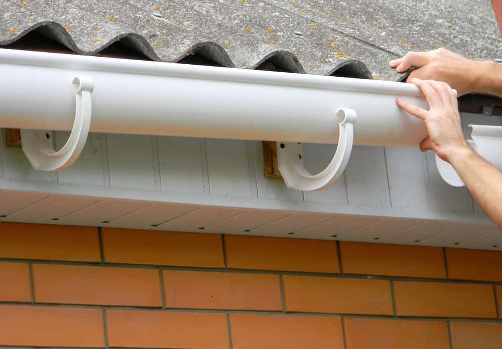 Close-up of hands installing a white plastic rain gutter on the edge of a roof with grey shingles above a wall of orange bricks.