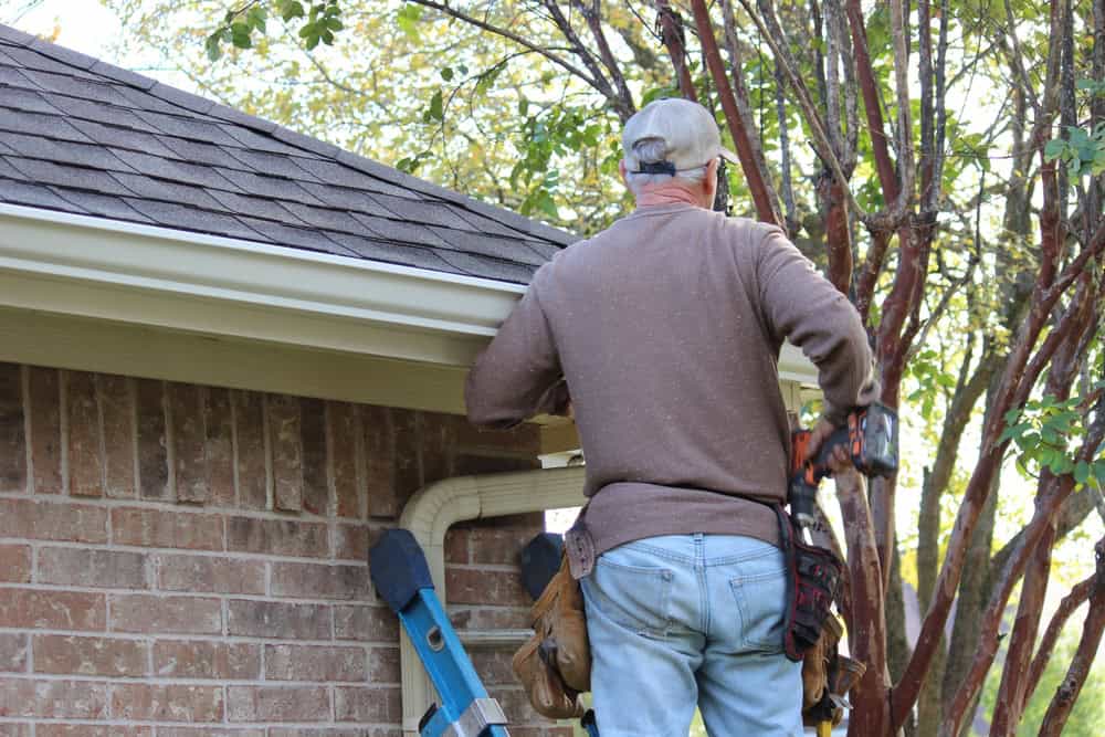 A man wearing a cap and toolbelt works on the gutter of a brick house, with a blue ladder leaning against the wall and trees in the background.