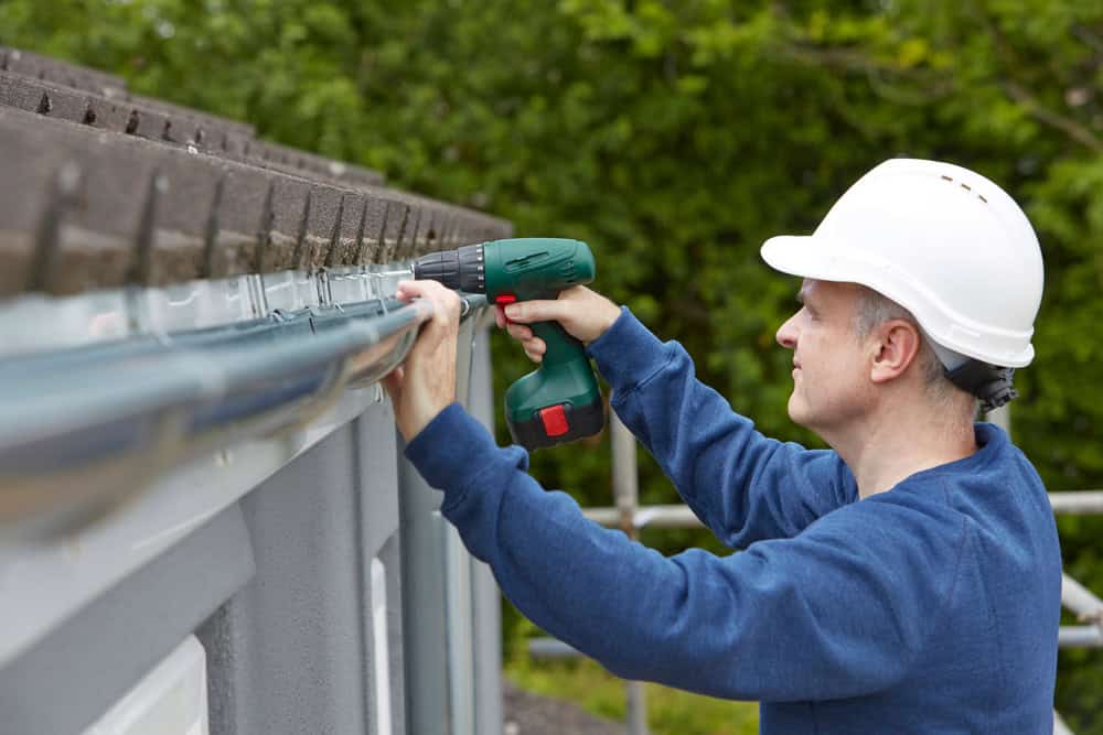 A construction worker in a white hard hat uses a cordless drill to install or repair a gutter on the edge of a building roof, with trees and greenery in the background.