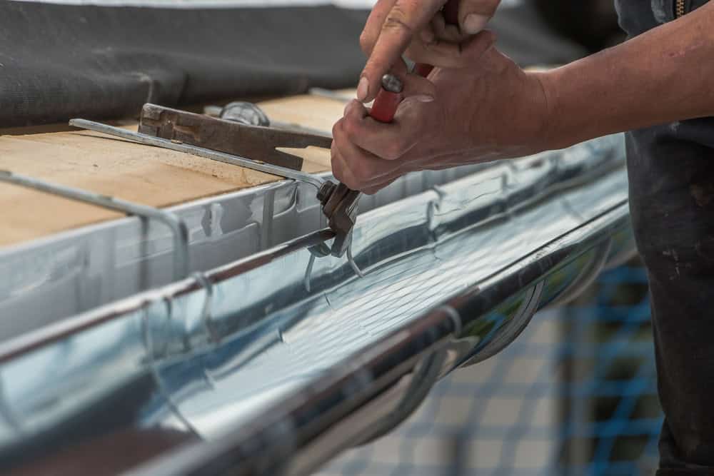 A person uses pliers and holds metal while installing or repairing a metal gutter on a building, with wooden boards and a netted safety barrier visible in the background.
