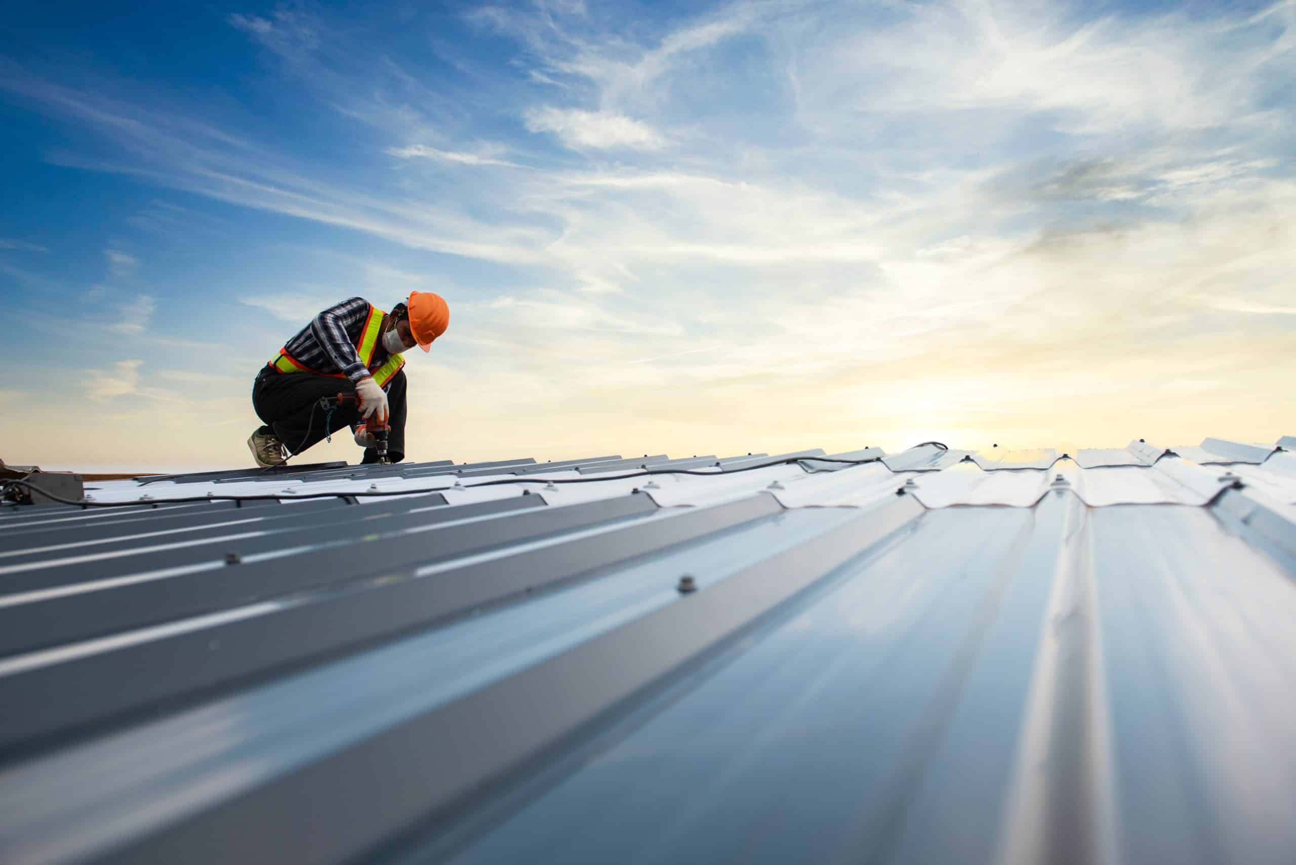 A construction worker in safety gear kneels on a metal roof, using a power tool to install or repair the panels under a bright, partly cloudy sky.